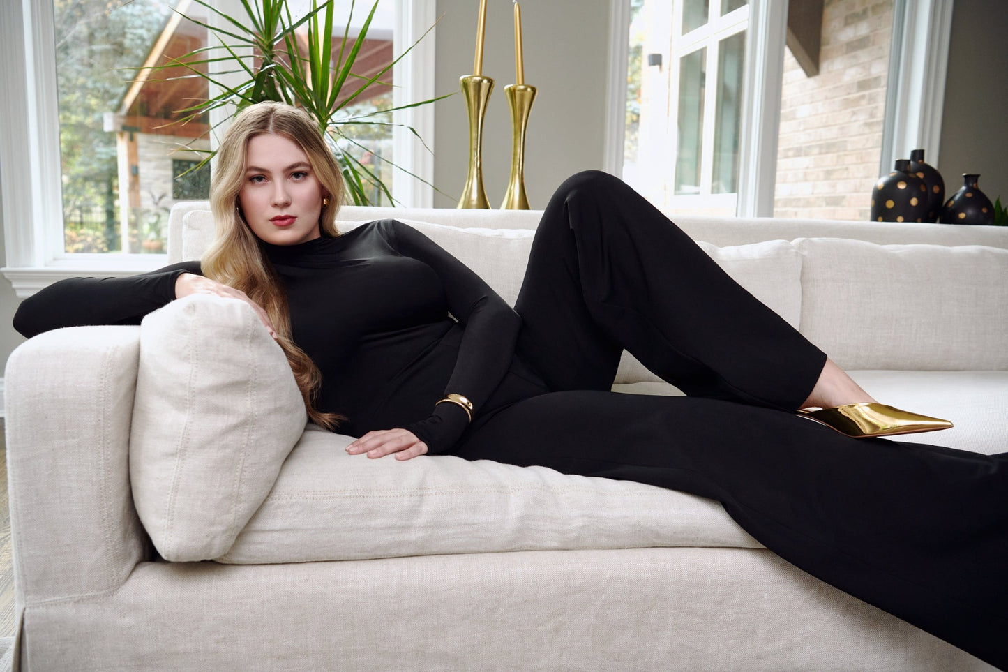Woman lounging on a sofa wearing a black stretch viscose fuller bust long sleeve mock neck pullover by Miriam Baker.