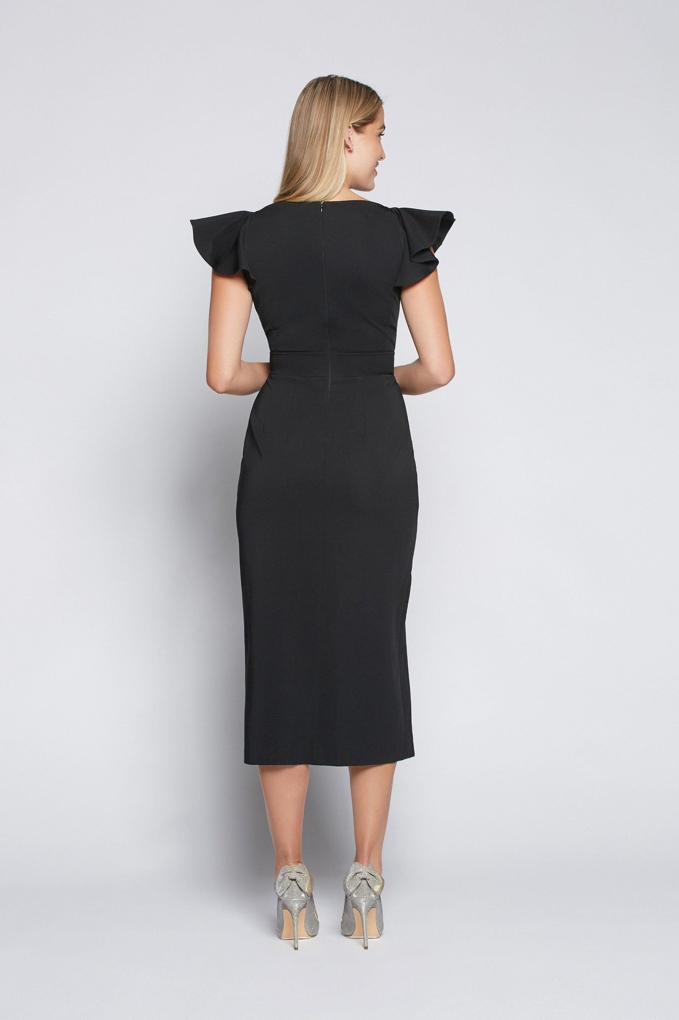 Back view of a woman wearing a black stretch viscose fuller bust boatneck midi dress with front slit, waist sash, and flutter sleeves designed by Miriam Baker.