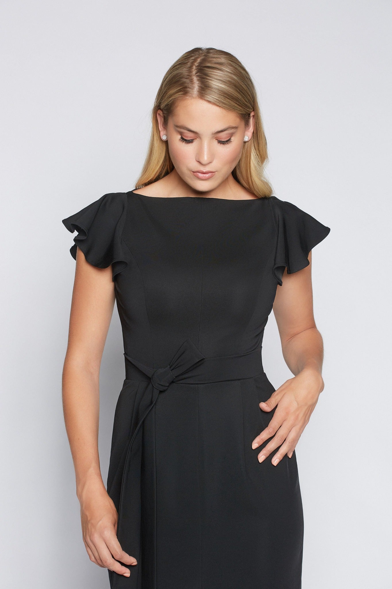 Front view of a woman wearing a black stretch viscose fuller bust boatneck midi dress with front slit, waist sash, and flutter sleeves designed by Miriam Baker.