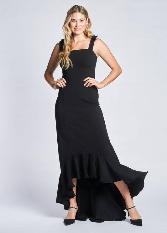 Front view of a woman wearing a black stretch viscose fuller bust empire waist gown with high low flounce hem and shoulder bow detail designed by Miriam Baker.