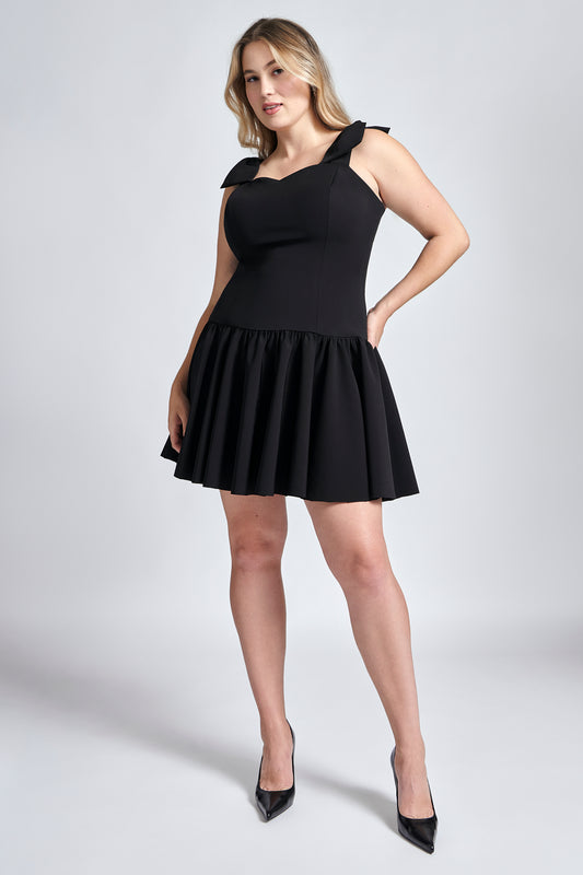 Front view of a woman wearing a black stretch viscose fuller bust dropped waist dress with full skirt and shoulder bows designed by Miriam Baker.