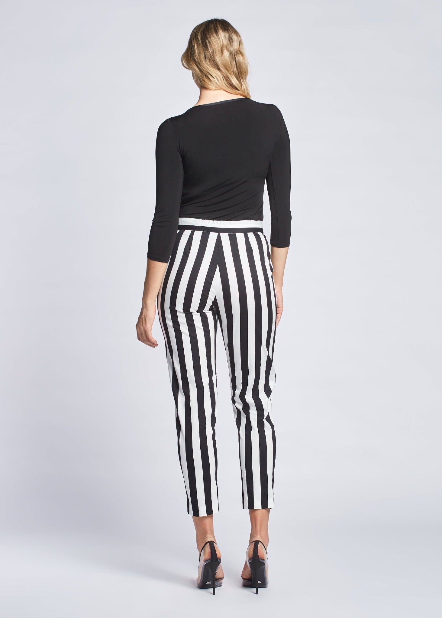 Back view of a woman wearing a pair of black and white stripe crop pants with slash pockets, a fly front, and drawstring waist designed by Miriam Baker.