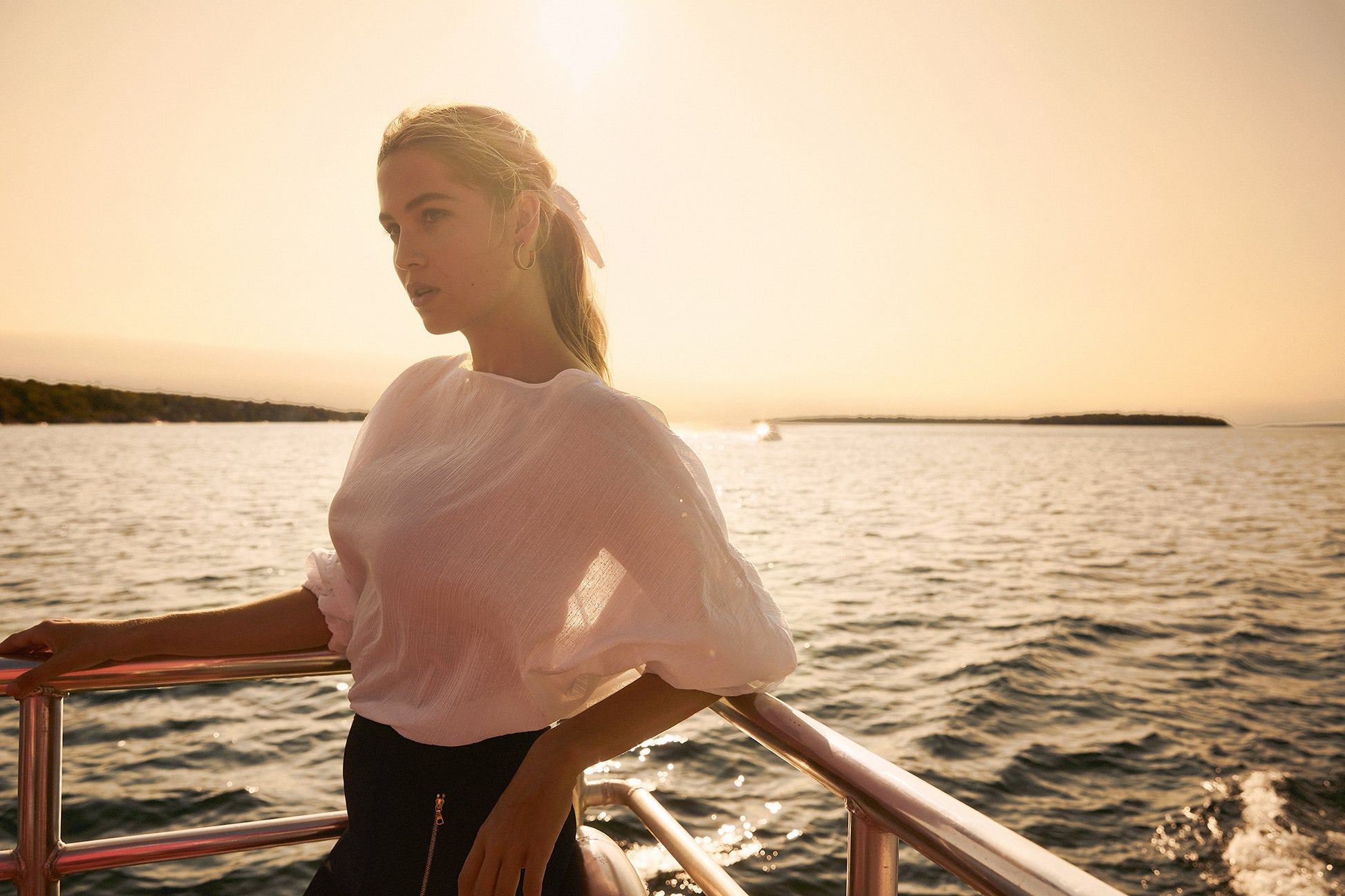 Woman on a boat at sunset wearing a cream coloured lurex linen boatneck fuller bust blouse with wrist ruffles and an elastic waist designed by Miriam Baker.
