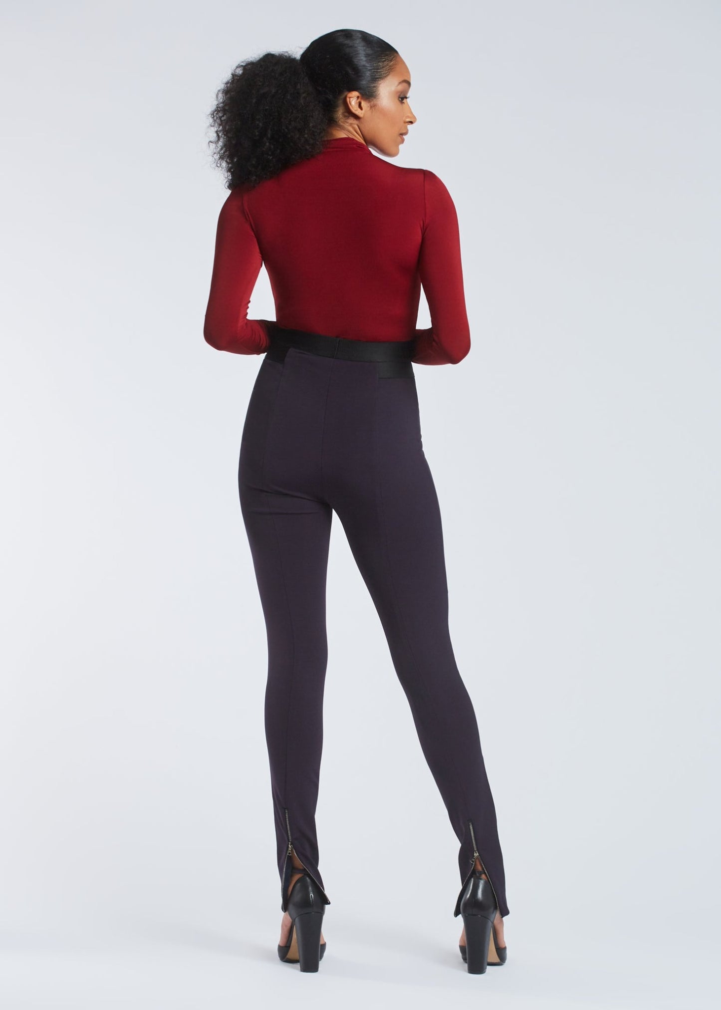 Back view of a woman wearing high waisted full length stretch trousers in navy blue with an exposed zipper at the hem designed by Miriam Baker.