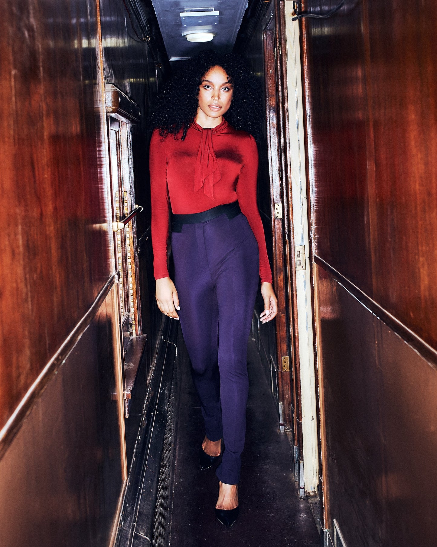 Front view of a woman walking in high waisted full length stretch trousers in navy blue with an exposed zipper at the back hem designed by Miriam Baker.