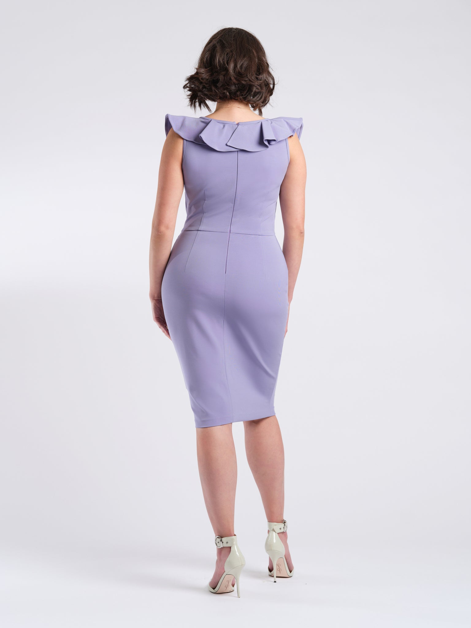 Back view of a woman wearing a stretch viscose fuller bust v-neck sheath dress with ruffles that frame the neckline in lilac designed by Miriam Baker.