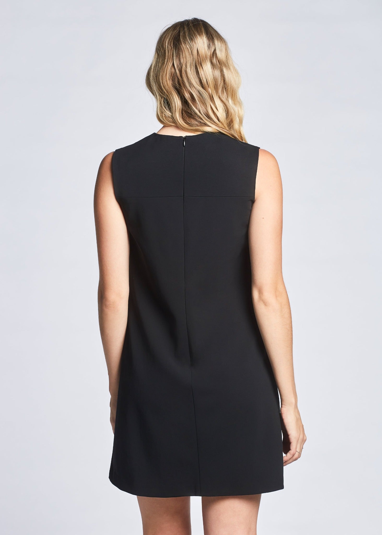 Back view of a woman wearing a black semi-fitted-fuller bust A-line shift dress with a crew neck and welt pockets designed by Miriam Baker.
