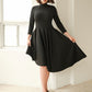 Front view of a woman wearing an asymmetric mock neck fuller bust dress with 3/4 length sleeves and side seam pockets in black stretch viscose fabric by Miriam Baker.