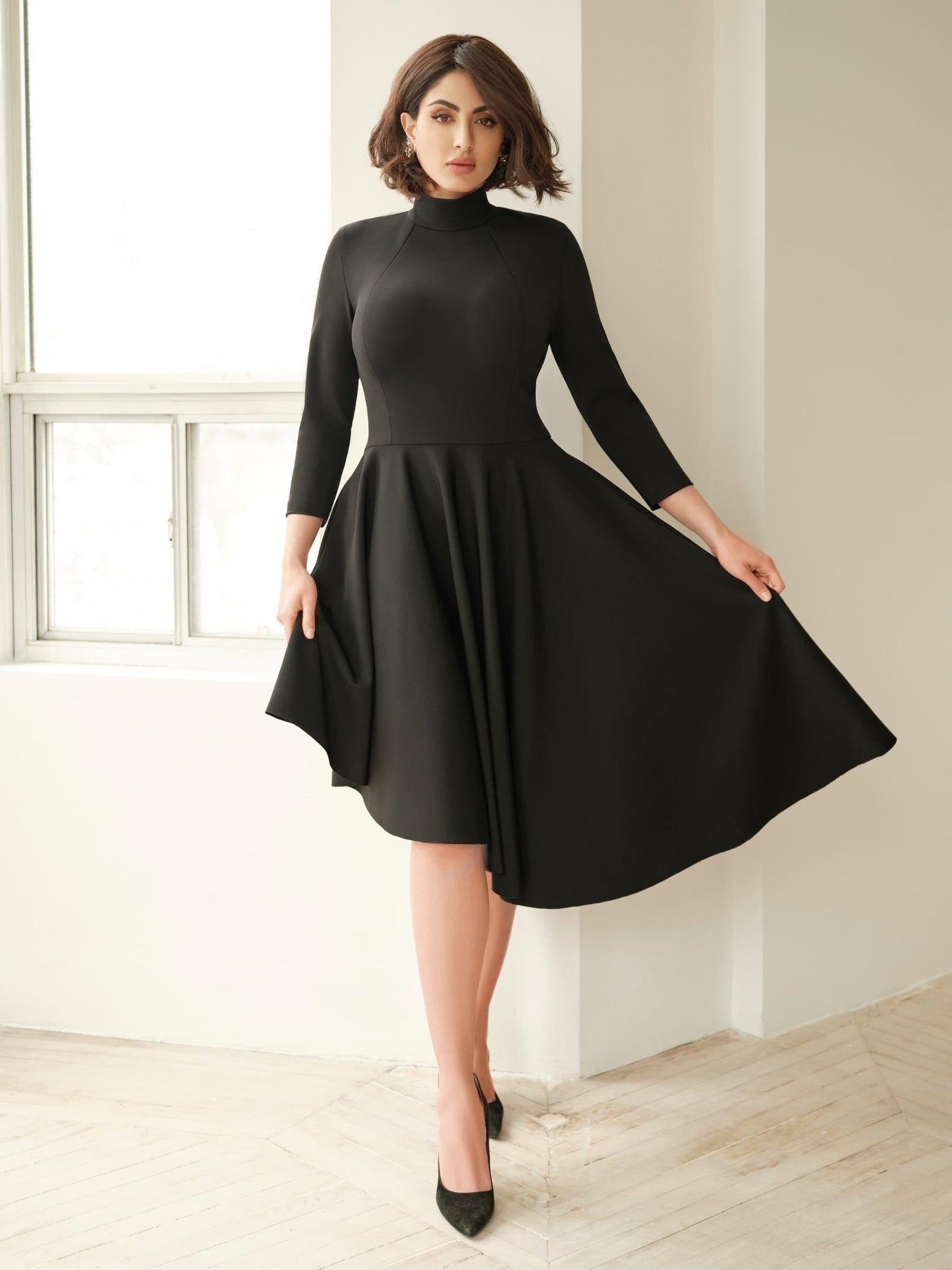 Front view of a woman wearing an asymmetric mock neck fuller bust dress with 3/4 length sleeves and side seam pockets in black stretch viscose fabric by Miriam Baker.