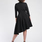 Back view of a woman wearing an asymmetric mock neck fuller bust dress with 3/4 length sleeves and side seam pockets in black stretch viscose fabric by Miriam Baker.