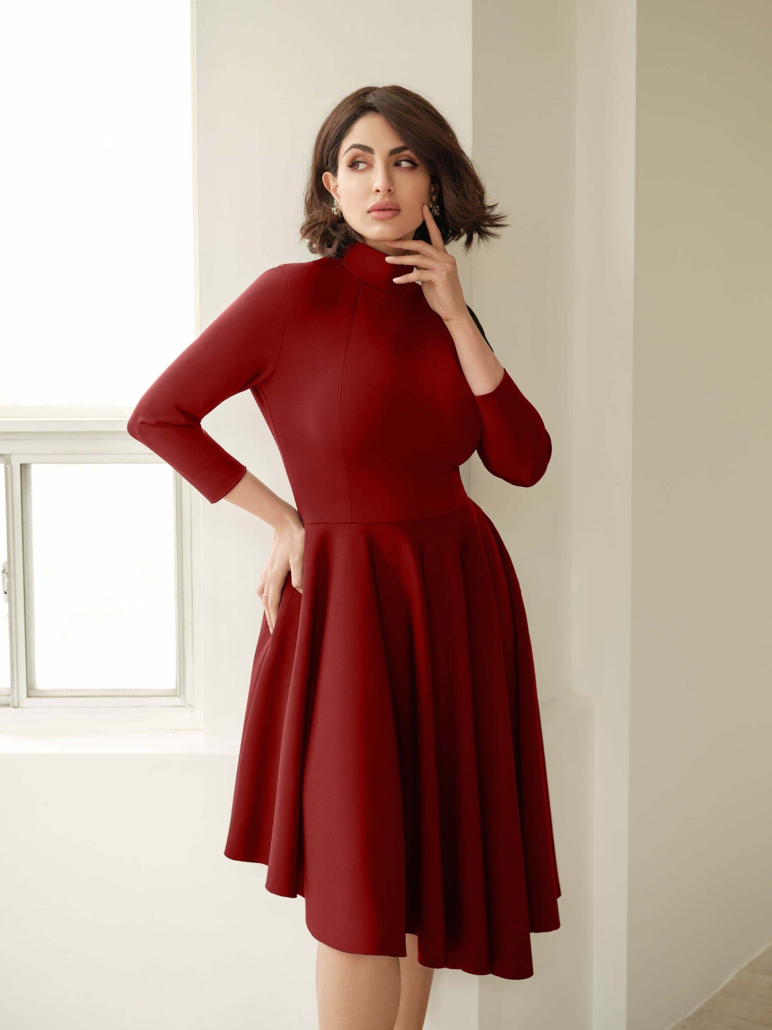Front view of a woman wearing an asymmetric mock neck fuller bust dress with 3/4 length sleeves and side seam pockets in red stretch viscose fabric by Miriam Baker.