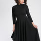 Front view of a woman wearing a asymmetric mock neck fuller bust dress with 3/4 length sleeves and side seam pockets in black stretch viscose fabric by Miriam Baker.