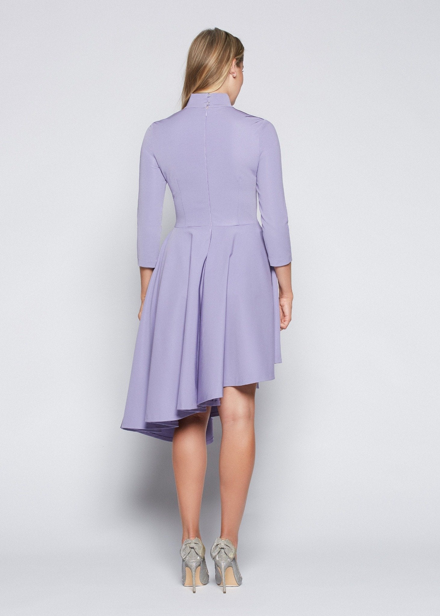 Back view of a woman wearing an asymmetric mock neck fuller bust dress with 3/4 length sleeves and side seam pockets in lilac stretch viscose fabric by Miriam Baker.