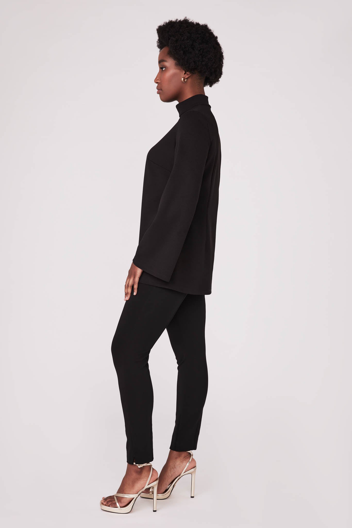 Side view of a woman wearing a black fuller bust tunic style top with flared sleeves in stretch crepe fabric designed by Miriam Baker.