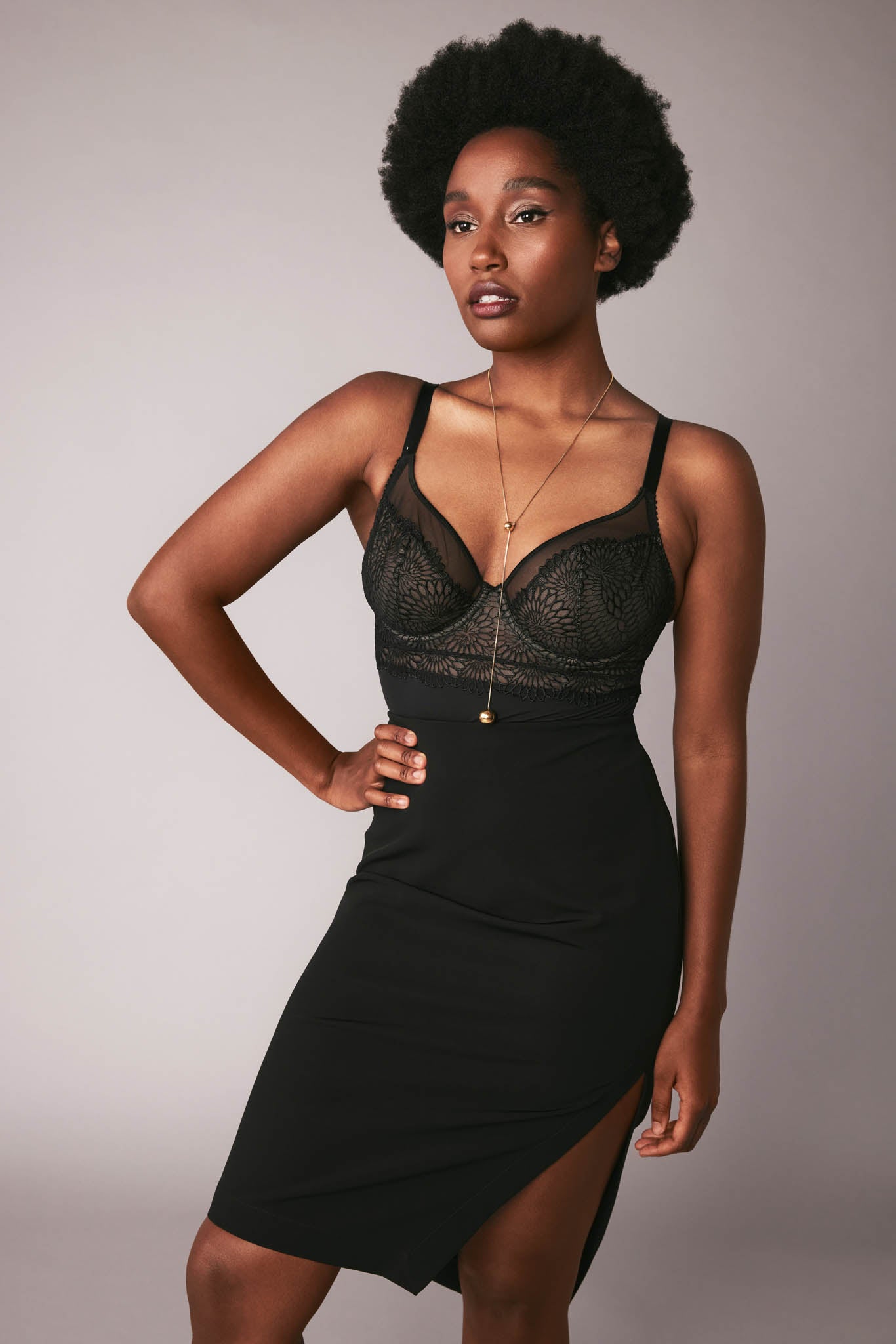 Front view of a woman wearing a black DD+ open back bodysuit with built in full support underwire bra paired with a black stretch viscose asymmetric skirt by Miriam Baker. 